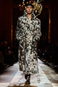 Model on a runway wearing a long black-and-white spotted faux fur coat with a black headband under chandeliers.