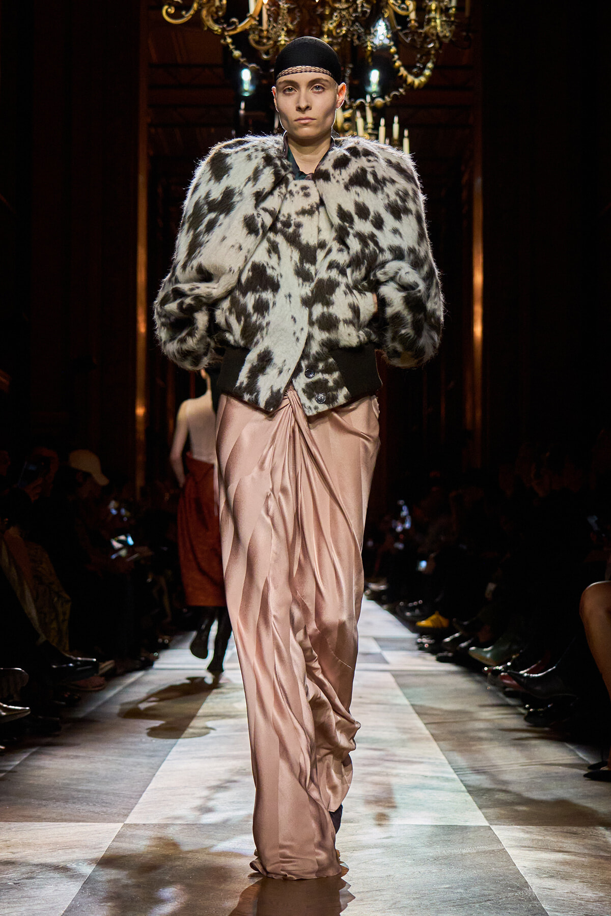 Model on a fashion runway wearing a black-and-white spotted fur coat over a pale pink satin gown.