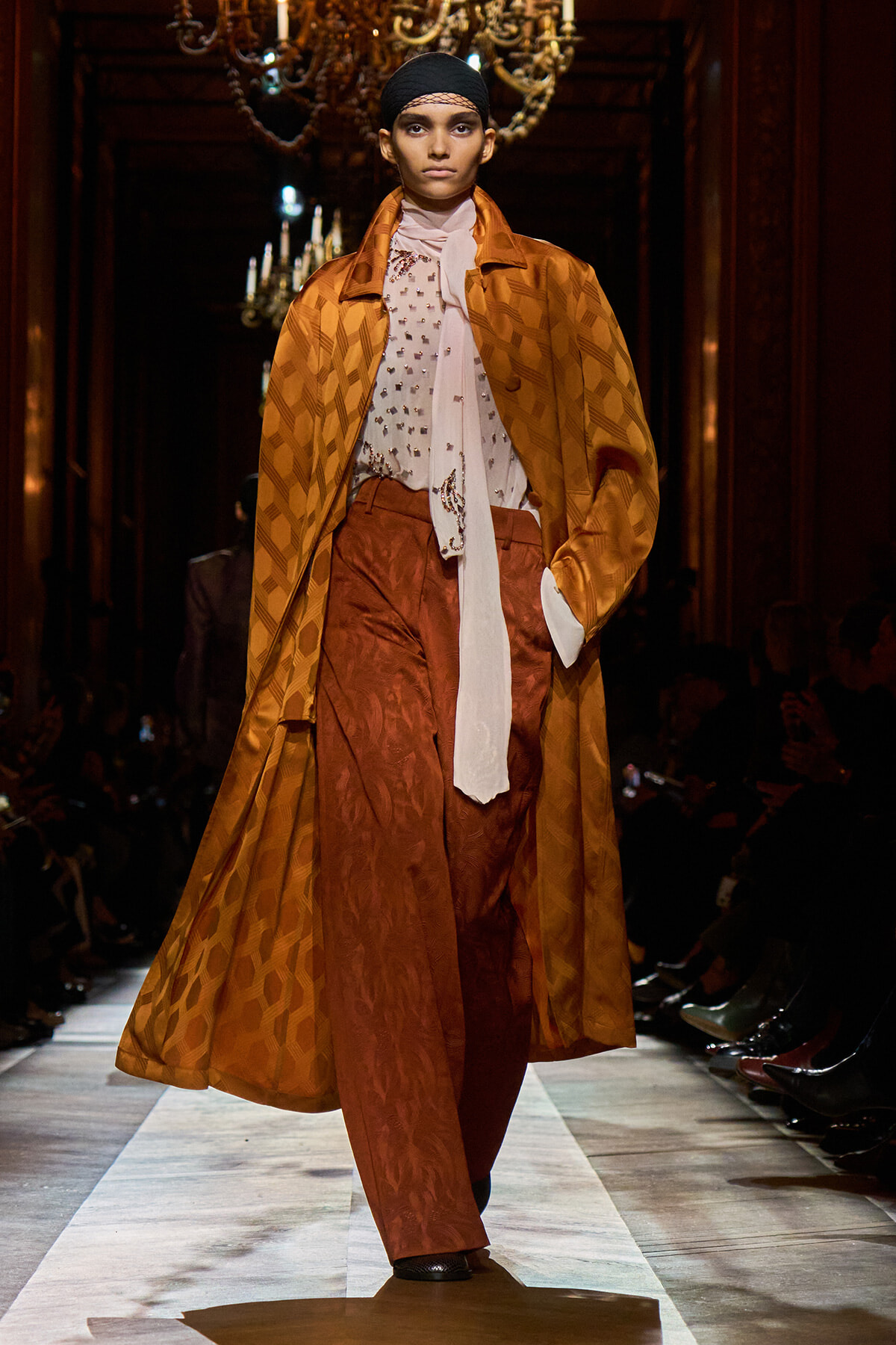 Model walking down a runway in an orange patterned coat over a rust-brown suit with a pale blouse and white scarf, under chandeliers in a dark venue.