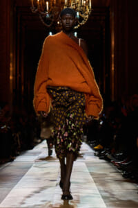 Model on a runway wearing an oversized orange knit sweater draped over one shoulder with a tiered, multicolored floral skirt and flat sandals, walking toward the camera.