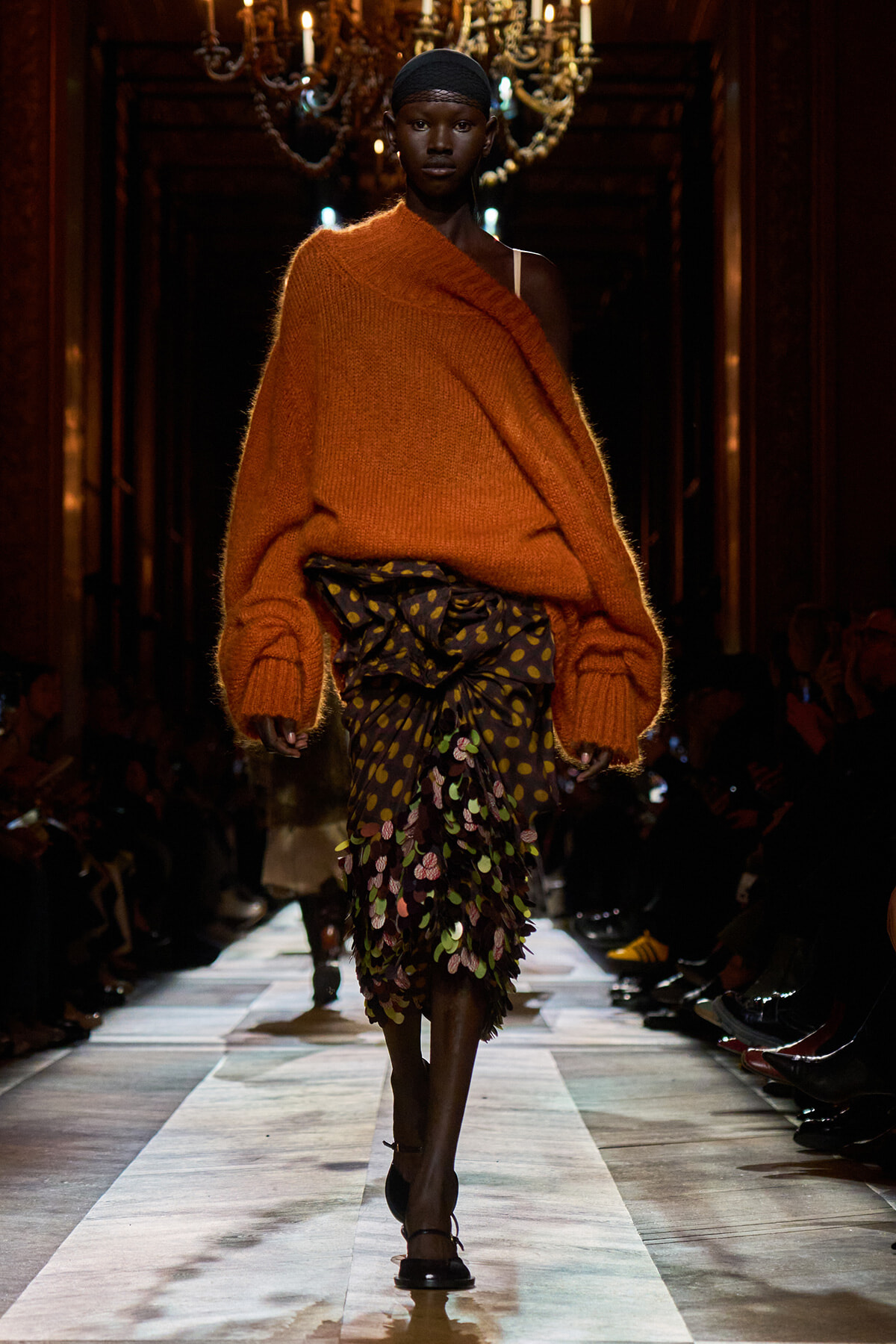 Model on a runway wearing an oversized orange knit sweater draped over one shoulder with a tiered, multicolored floral skirt and flat sandals, walking toward the camera.