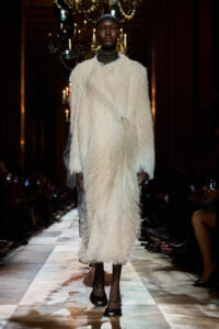 Model on a runway wearing a long white shaggy coat, chandeliers overhead in a dim fashion show.