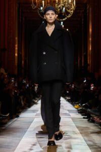 Model walking the runway in a loose black double-breasted coat with a headscarf, chandeliers overhead, audience on sides.