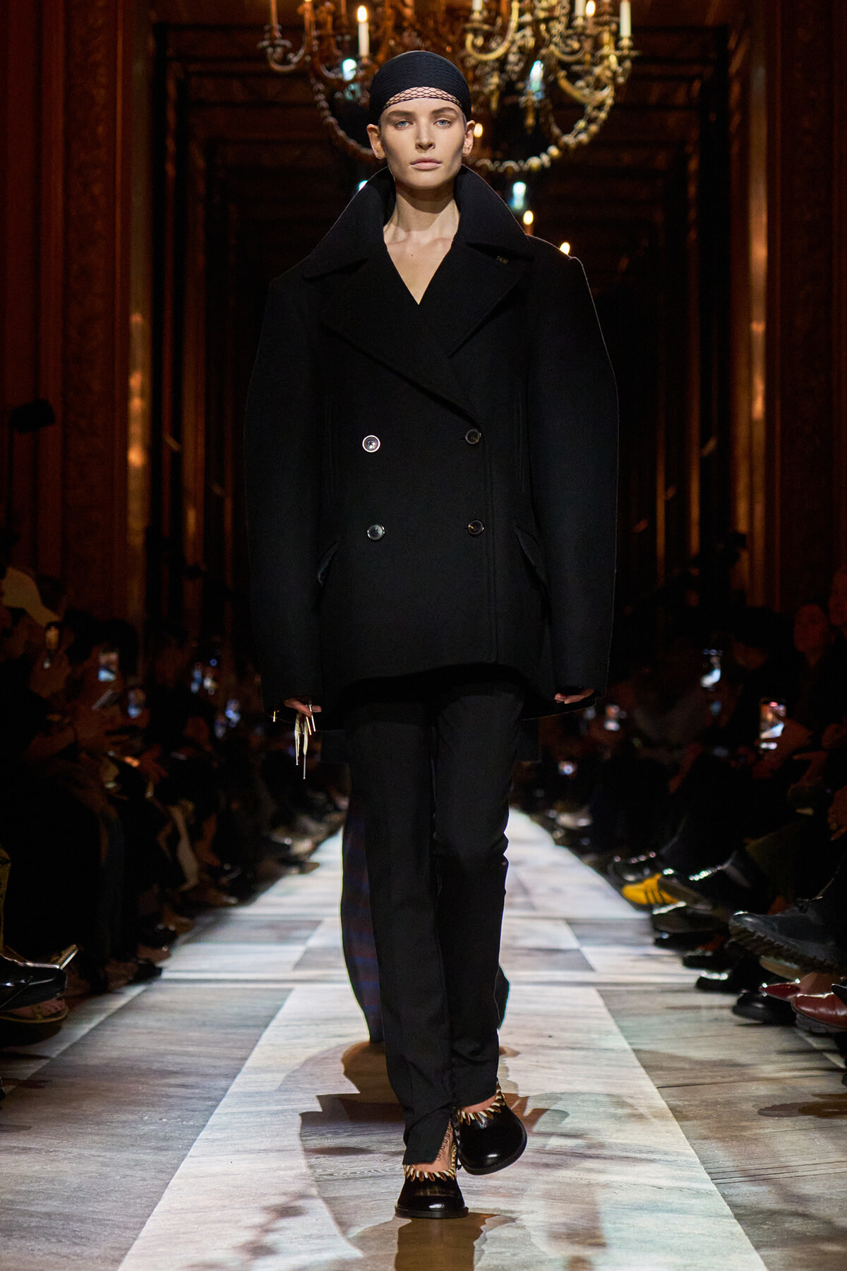 Model walking the runway in a loose black double-breasted coat with a headscarf, chandeliers overhead, audience on sides.