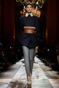 Model walks a fashion runway in a navy oversized blazer, cream collar, wide braided belt, gray trousers, and white shoes under chandeliers.