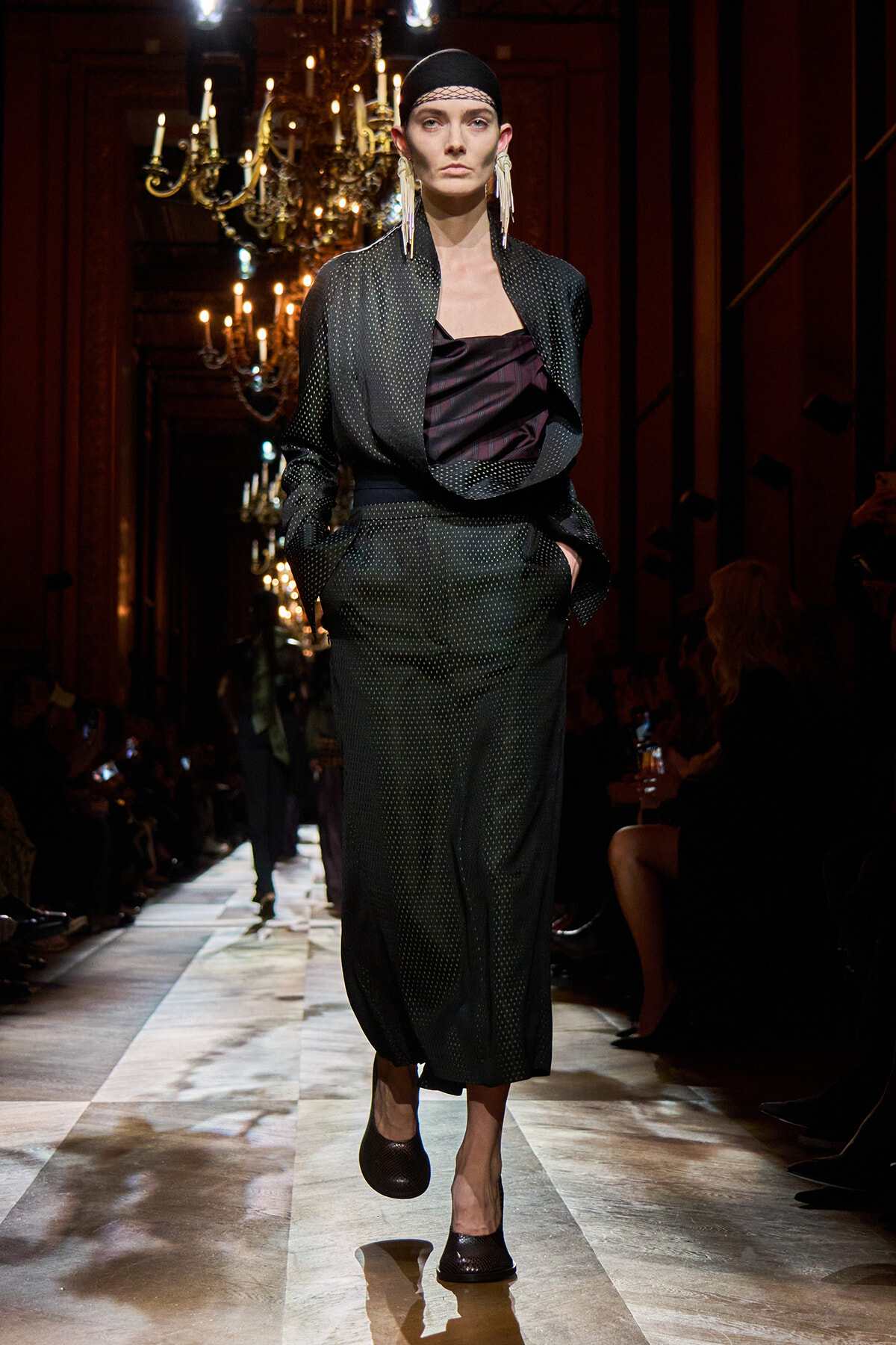 Female model walking a runway in a dark dotted two-piece suit with a draped satin top, headscarf, and long fringe earrings against a chandelier-lit backdrop.