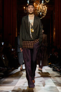 Model walking the fashion runway in a dark pinstriped blouse, patterned belt, layered white necklaces, and burgundy-striped wide-leg pants under chandelier lighting.