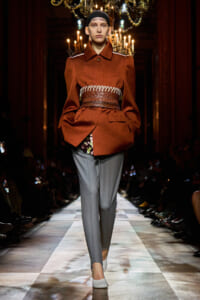 Model walks a runway in a rust-orange oversized coat cinched with a wide, textured belt, paired with gray trousers and white shoes under a chandelier-lit backdrop.