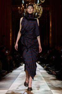 Model walking the runway in a dark blue and burgundy striped draped dress, chandelier backdrop.