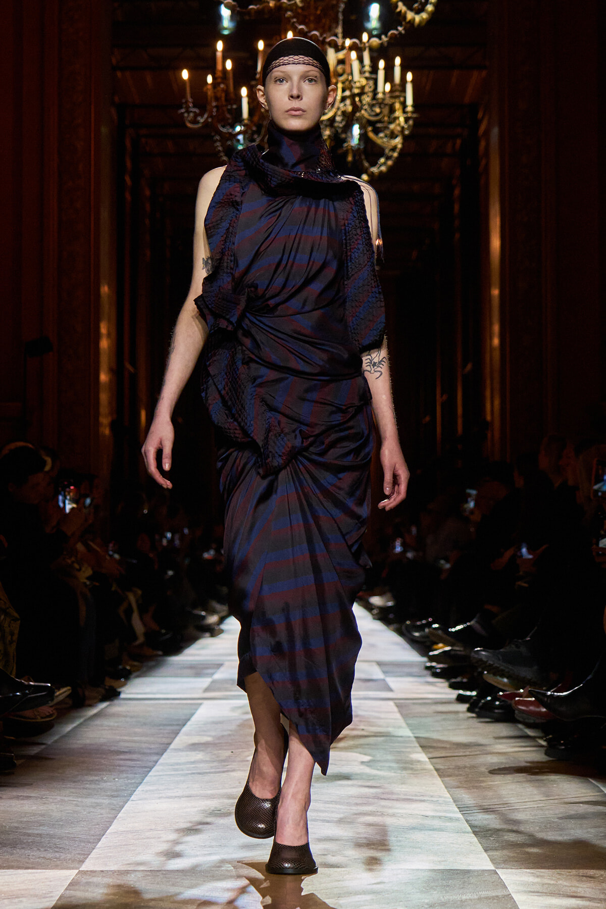 Model walking the runway in a dark blue and burgundy striped draped dress, chandelier backdrop.