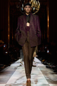 Model walking runway in a oversized burgundy paisley suit with matching pants, black headband, ornate chandelier backdrop