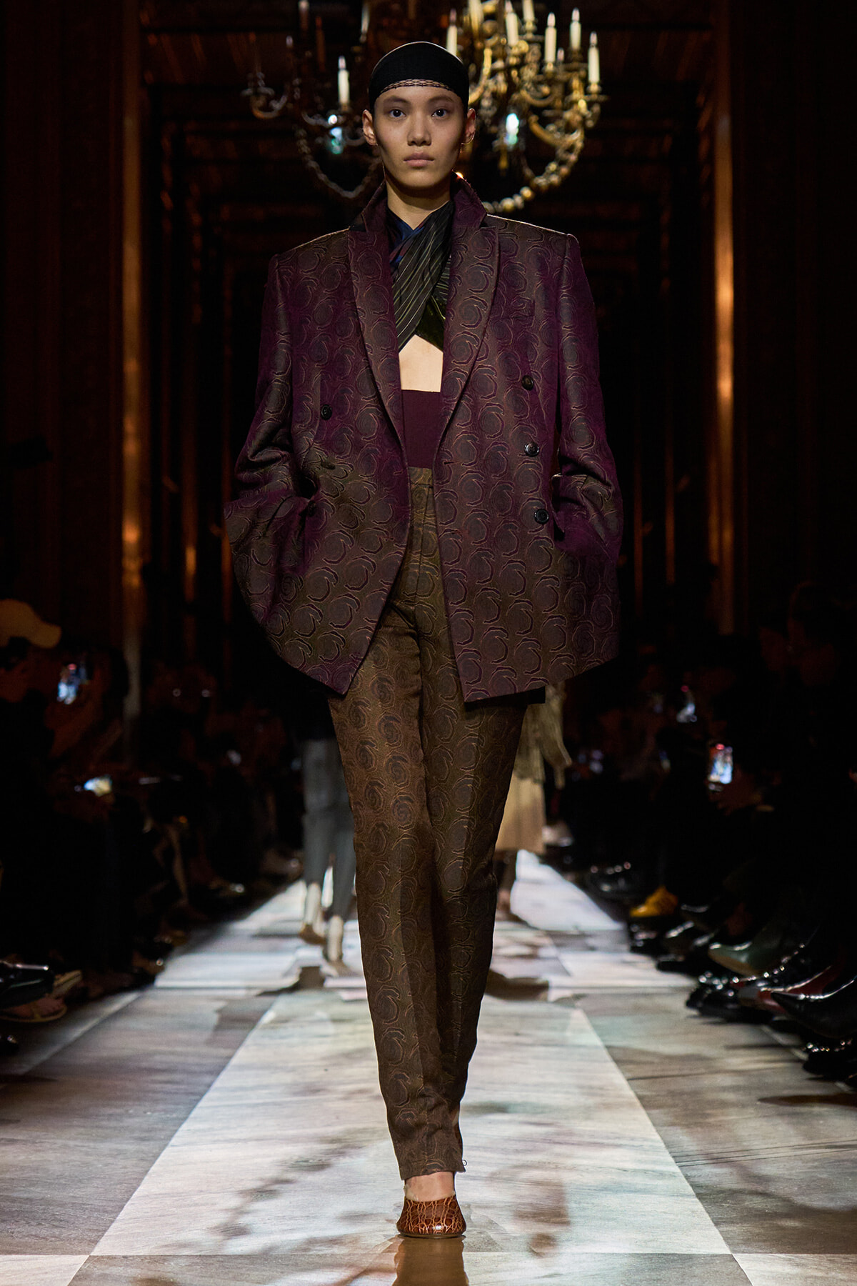 Model walking runway in a oversized burgundy paisley suit with matching pants, black headband, ornate chandelier backdrop