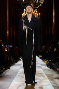 Model on a fashion show runway wearing a long black coat with white fringe detailing.