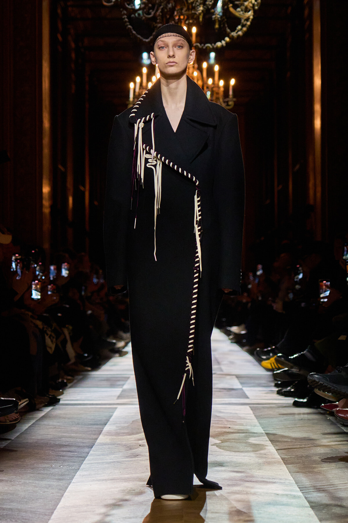Model on a fashion show runway wearing a long black coat with white fringe detailing.