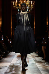 Model walks a fashion runway in a black, structured dress with a long fringe necklace; chandeliers glow overhead in a dim, elegant venue.