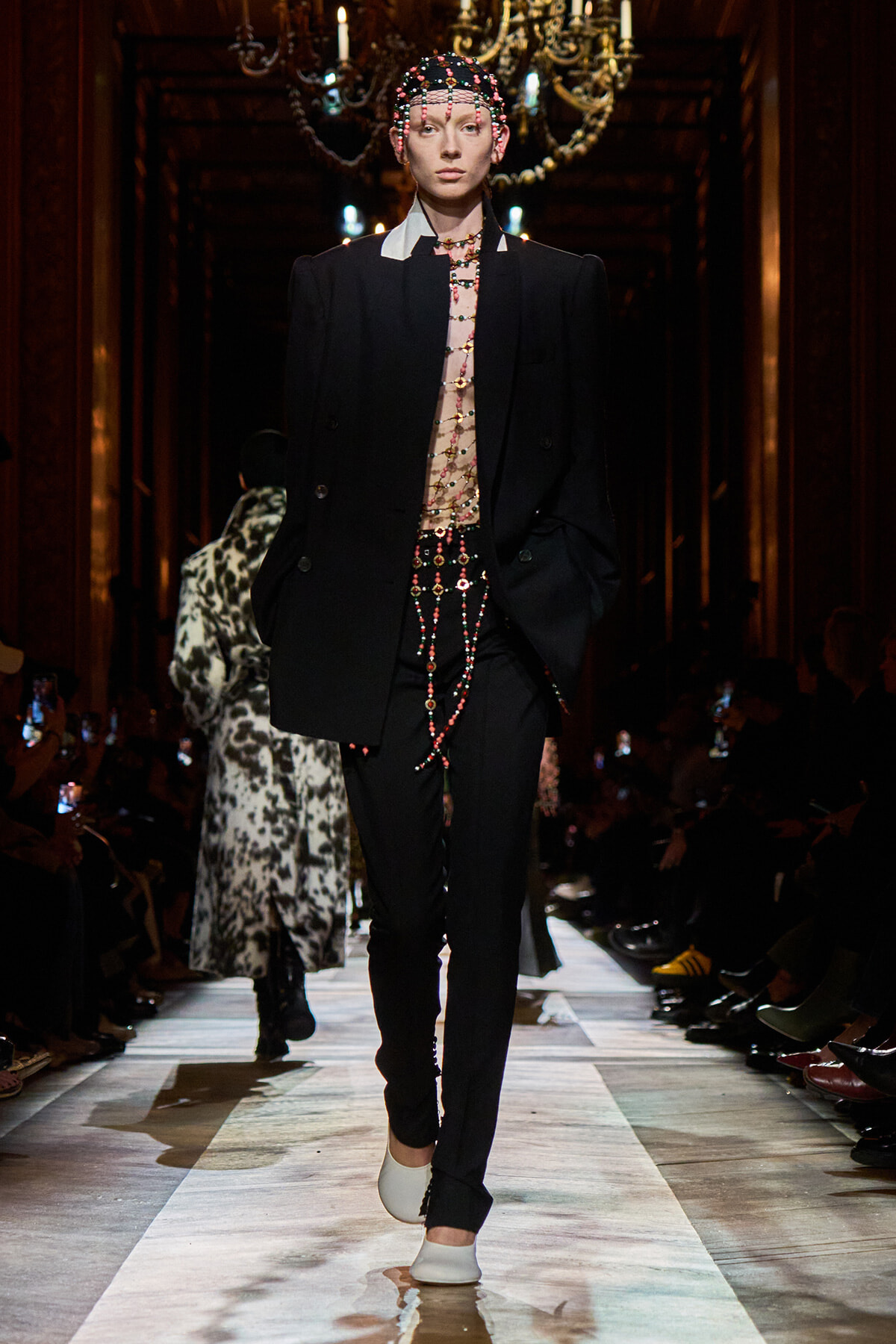 Model walking a runway in an oversized black suit with a sheer beaded torso, layered bead jewelry, and a beaded headpiece; white shoes and chandeliers in background.