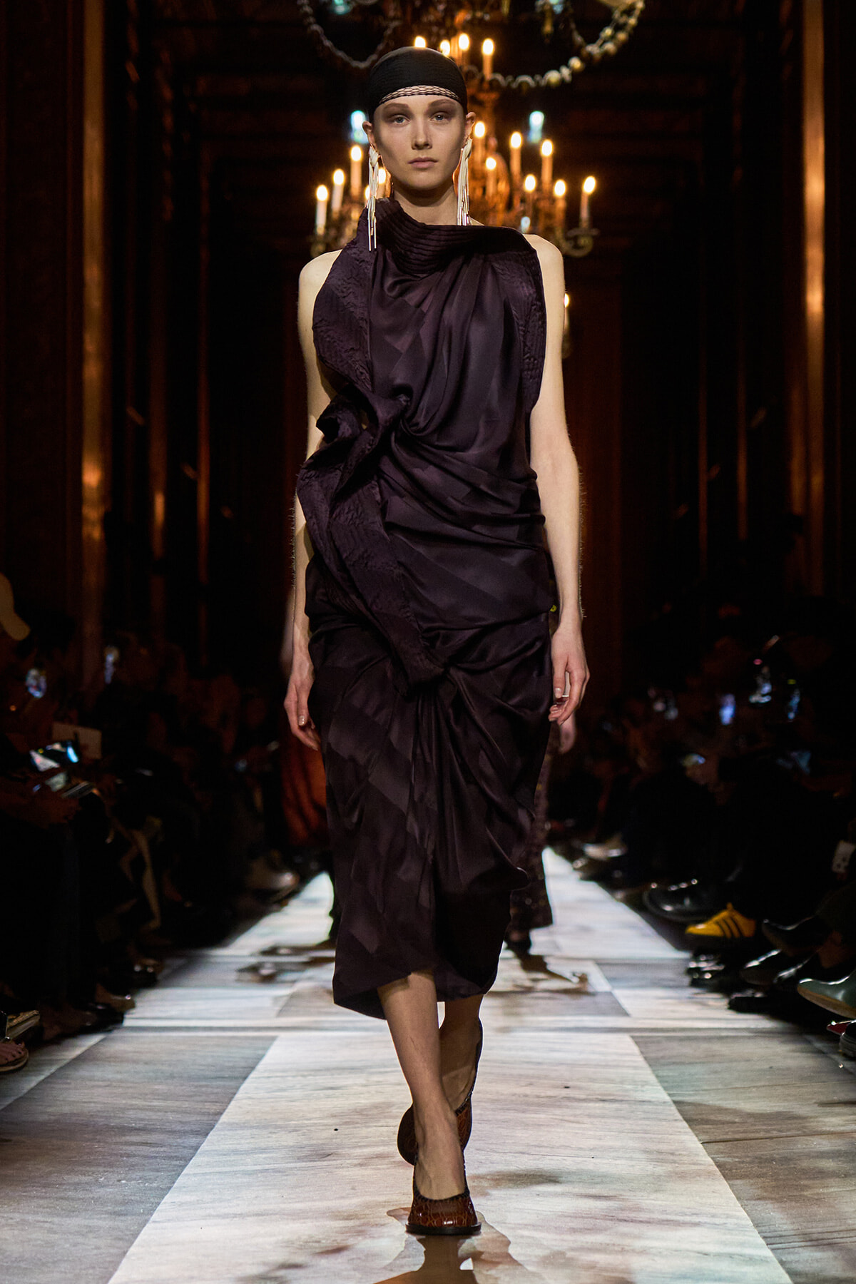 Model walks a runway in a dark purple draped gown, with a black headband and long earrings, chandeliers glowing in the background.
