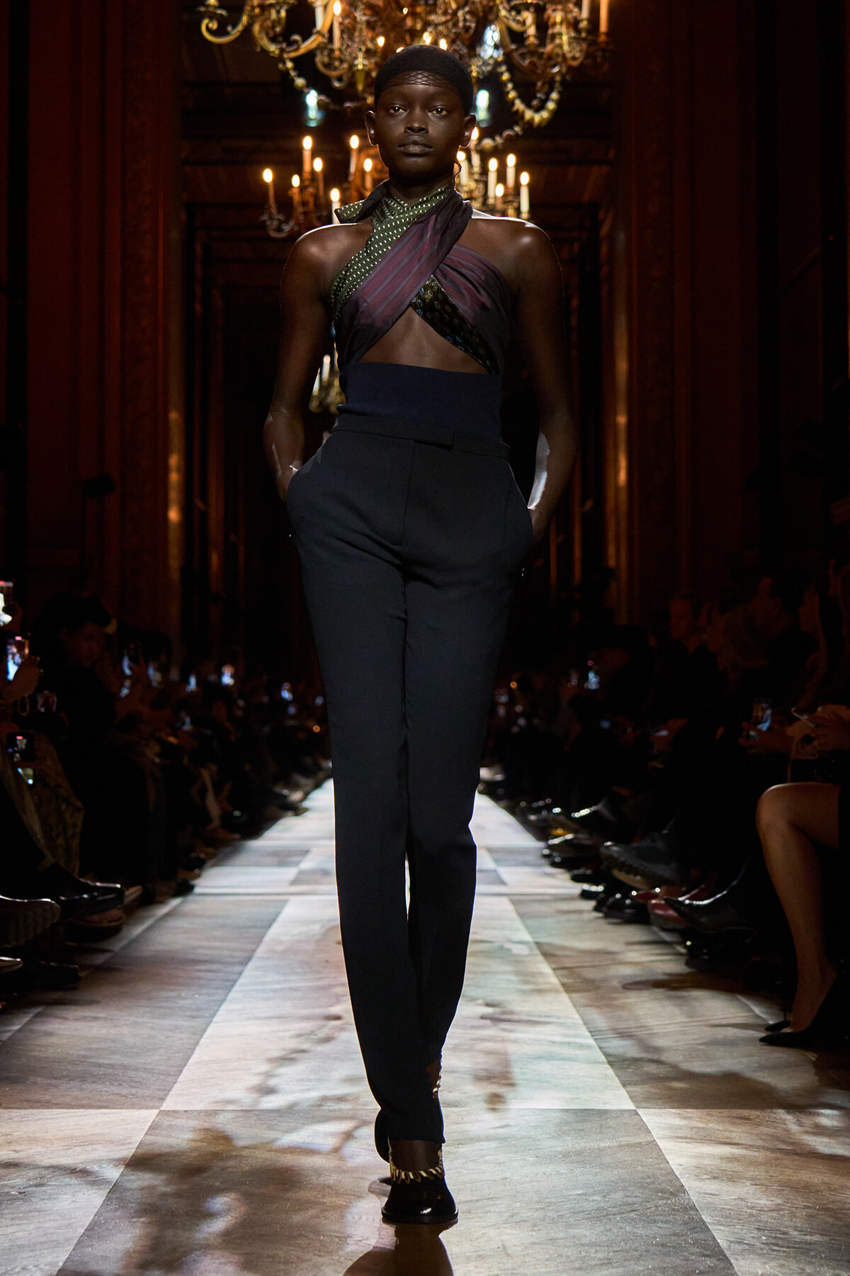 Model walking a fashion runway in a dark, chandeliers-lit hall, wearing a crisscross halter top and high-waisted navy trousers.