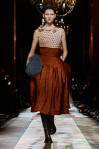 Model walks a runway in a beige sleeveless beaded top and rust-orange full skirt, holding a blue fluffy clutch under chandeliers.