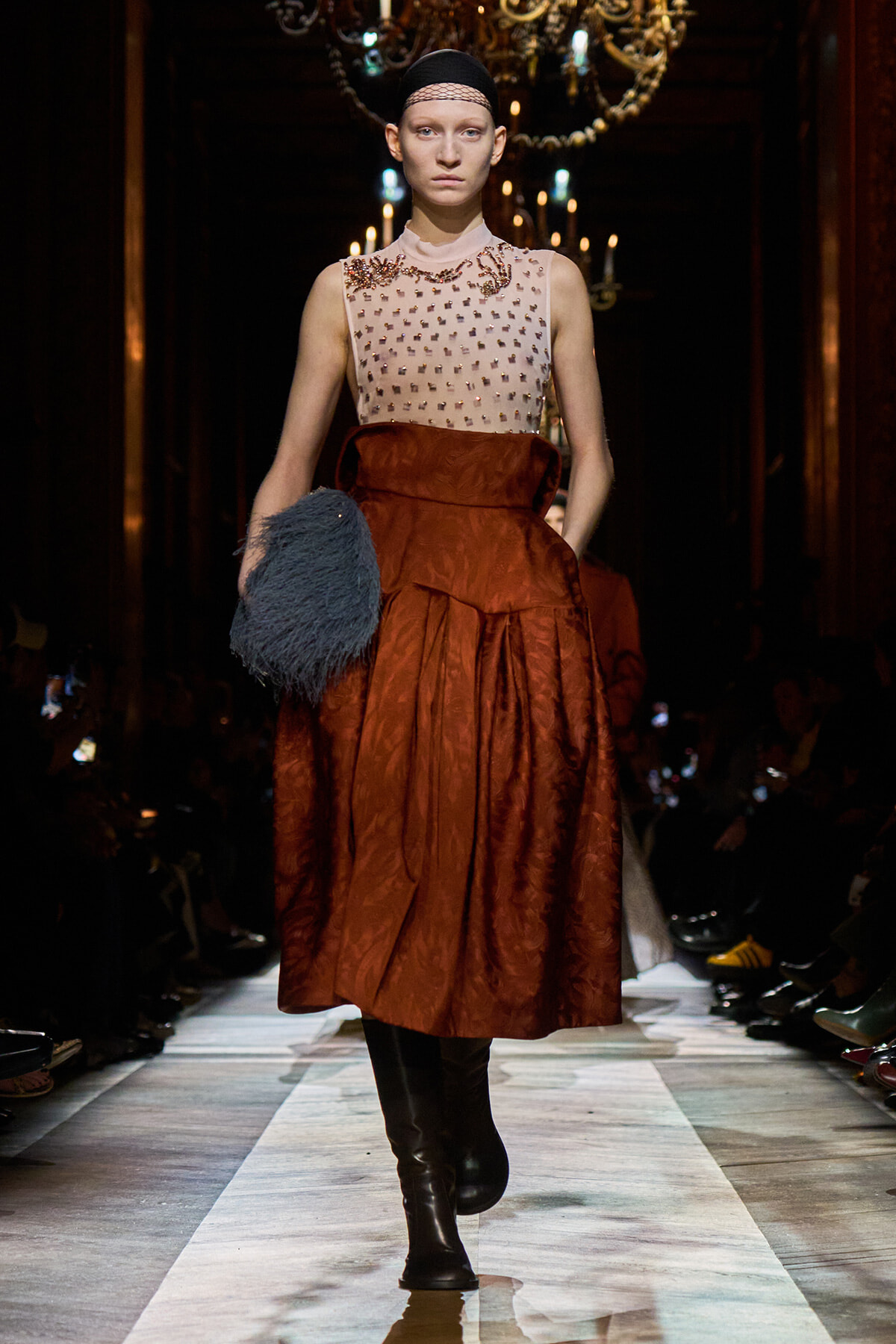Model walks a runway in a beige sleeveless beaded top and rust-orange full skirt, holding a blue fluffy clutch under chandeliers.