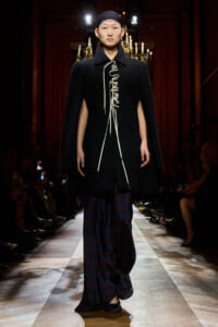Model walks a runway in a black coat with white lace-up detailing over dark striped pants.