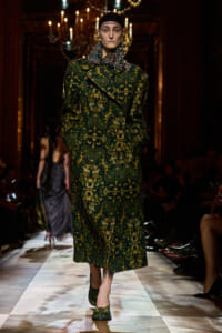 Model walks the runway in a long green brocade coat with a bold ornate pattern and matching green shoes, chandelier-lit backdrop behind.