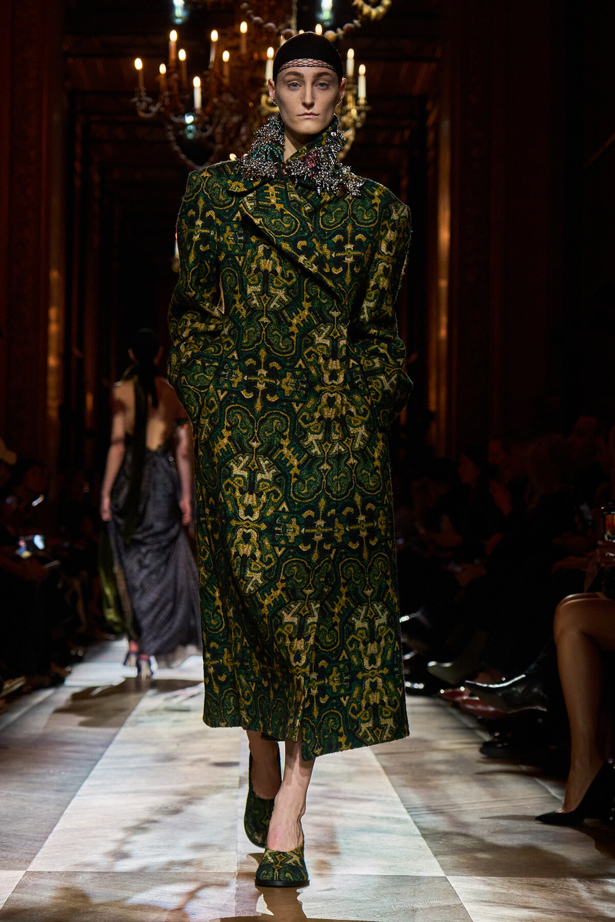 Model walks the runway in a long green brocade coat with a bold ornate pattern and matching green shoes, chandelier-lit backdrop behind.