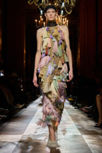 Model walking a runway in a multicolored tassel fringe gown with a black headband, chandeliers overhead.