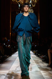 Model walks runway in a dark blue oversized cape-style top and teal patterned wide-leg pants, under chandeliers.