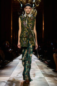 Model on runway wearing a green embroidered sleeveless vest with jewel embellishments over a pale shirt, paired with sheer green plaid pants and a black headband, walking forward confidently.