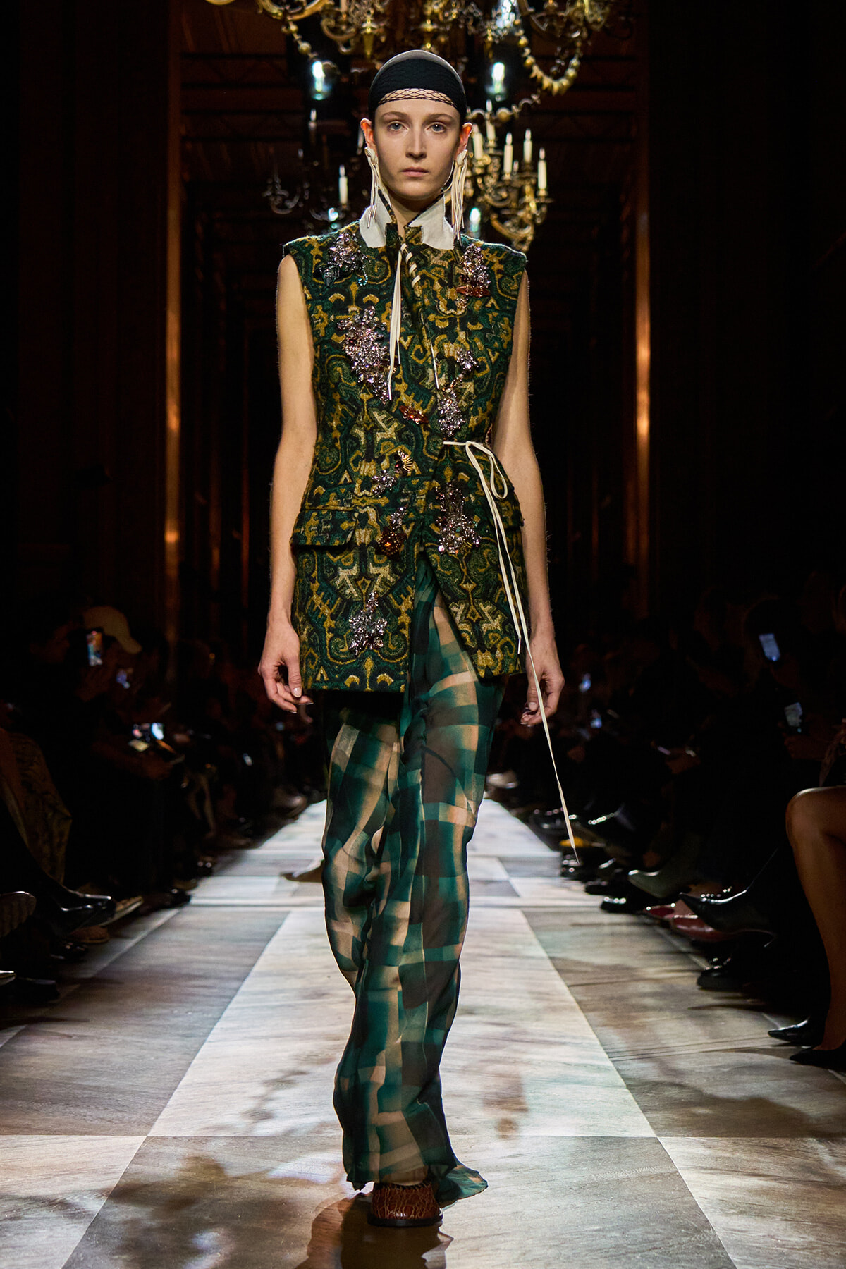 Model on runway wearing a green embroidered sleeveless vest with jewel embellishments over a pale shirt, paired with sheer green plaid pants and a black headband, walking forward confidently.