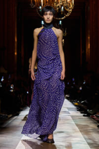 Model walks the runway in a flowing purple halter gown with white polka dots, under a chandelier-lit venue.