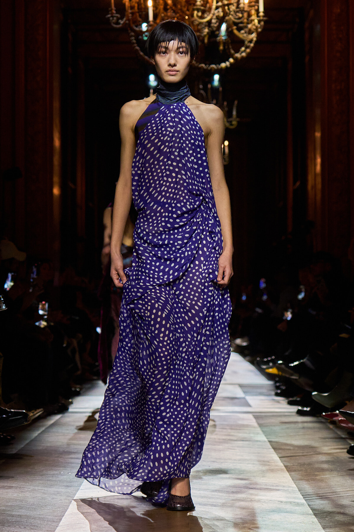 Model walks the runway in a flowing purple halter gown with white polka dots, under a chandelier-lit venue.