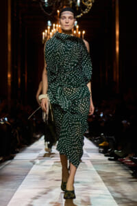 Model walks the runway in a dark venue wearing a green polka-dot, draped dress with a head wrap and black tights/shoes.