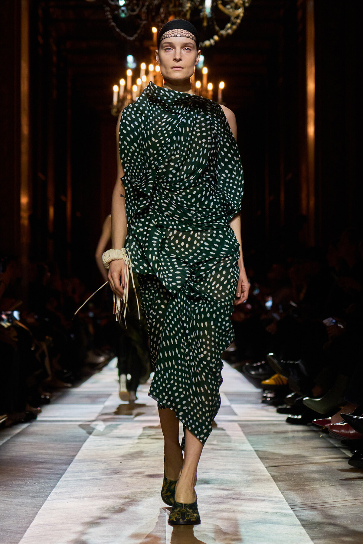 Model walks the runway in a dark venue wearing a green polka-dot, draped dress with a head wrap and black tights/shoes.