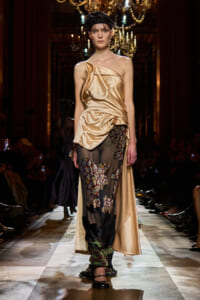 Model walks the runway in a one-shoulder champagne satin dress with a sheer, patterned underlay panel reaching to the floor.