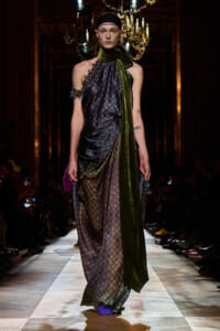 Model walking a fashion runway in a draped, patterned dress with a dark, glossy green scarf tied around the neck; ornate backdrop and chandeliers in a dimly lit venue.