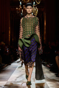 Model walking a runway in a green beaded sheer top with pink accents, a dramatic green sculptural waist and a striped purple skirt.