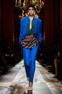 Model walking runway in a bright blue suit with a green blouse and patterned sash, chandeliers in the background.