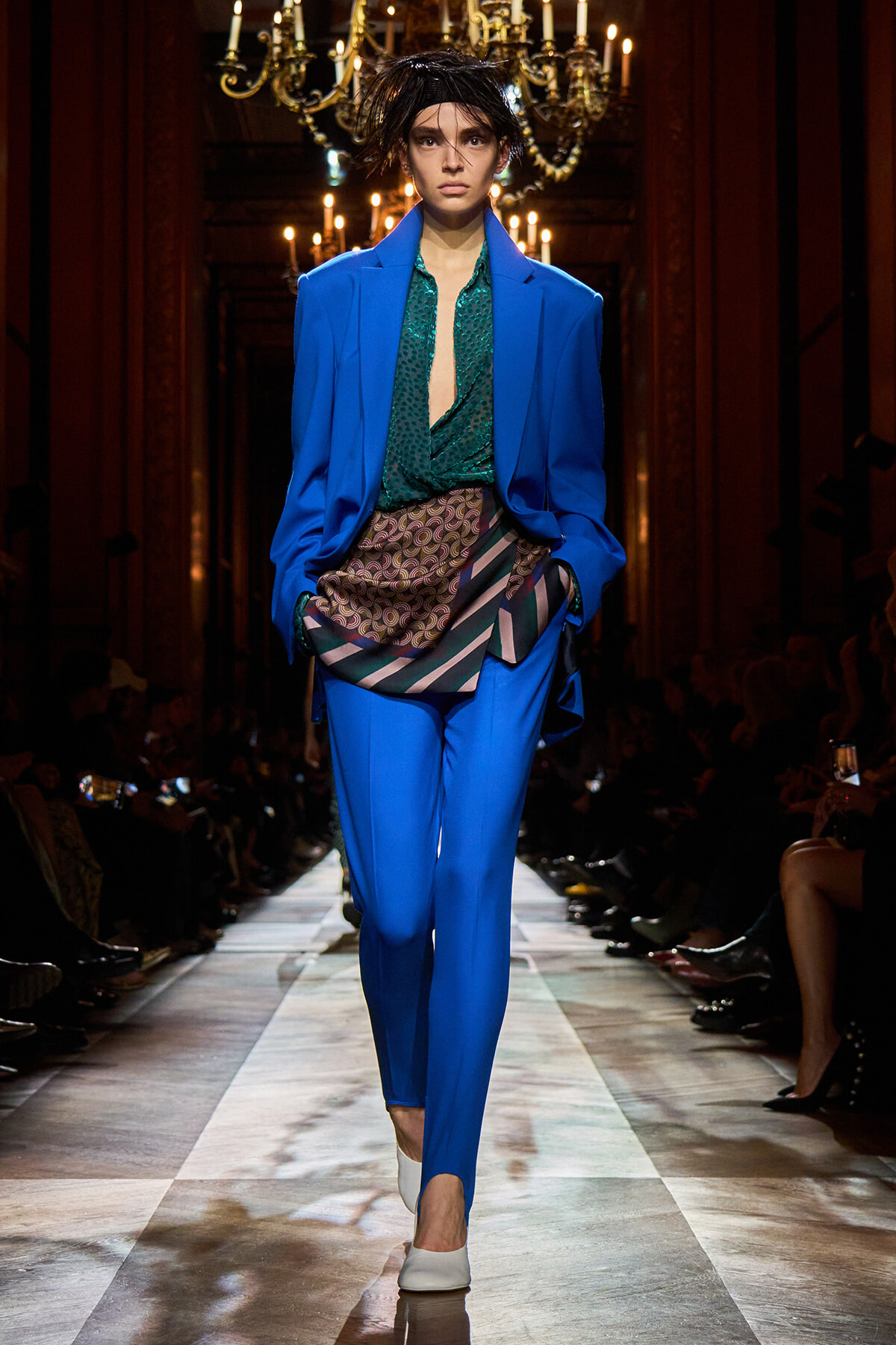 Model walking runway in a bright blue suit with a green blouse and patterned sash, chandeliers in the background.