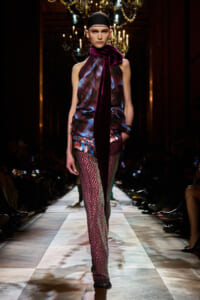 Model on runway wearing a burgundy-blue sheer top with a long velvet scarf draped around the neck and multicolored sequin-patterned leggings.