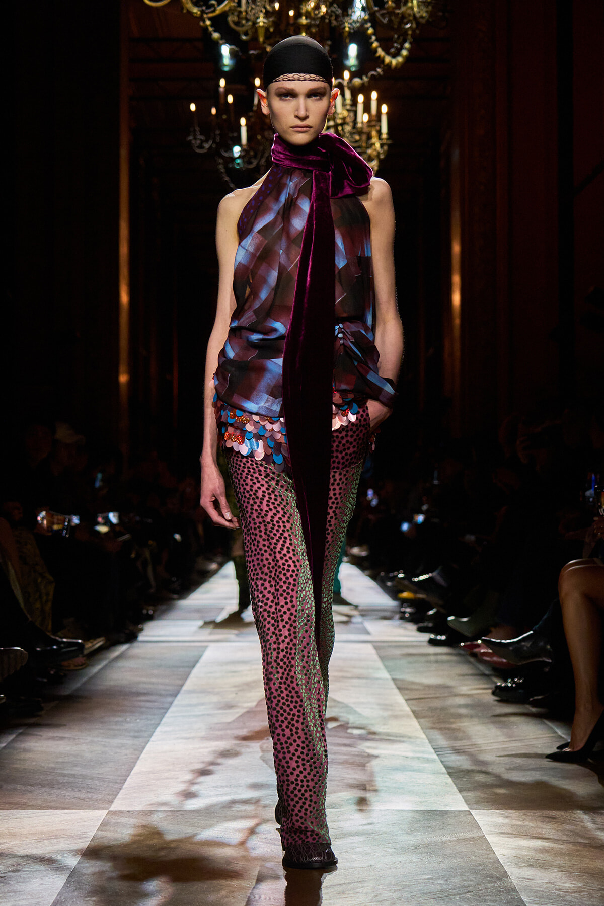 Model on runway wearing a burgundy-blue sheer top with a long velvet scarf draped around the neck and multicolored sequin-patterned leggings.