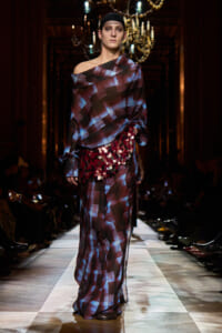 Model walks the runway in a burgundy-blue off-shoulder plaid gown with a sequined waist belt, chandeliers above in a dim, ornate hall.