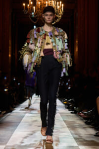 Model walks the runway in a multicolored tassel-fringe jacket over a burgundy top and black pants with side embellishments.