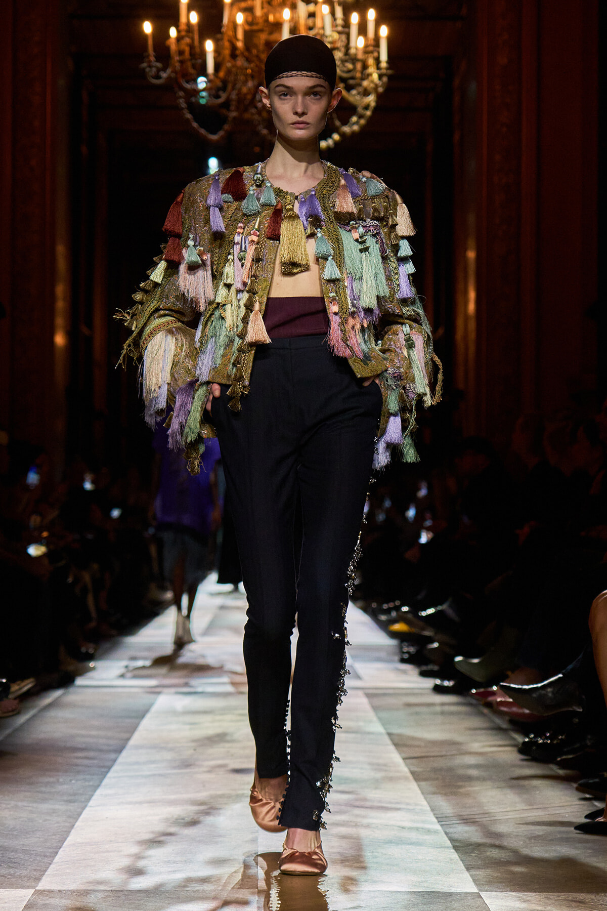 Model walks the runway in a multicolored tassel-fringe jacket over a burgundy top and black pants with side embellishments.