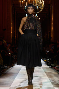 Model walks a fashion runway in a dark ornate hall wearing a high-waisted black voluminous skirt, patterned scarf top, and knee-high boots.