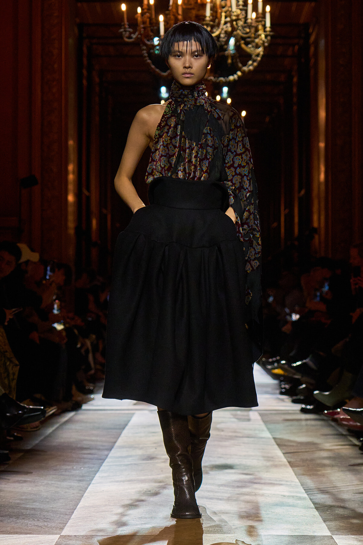 Model walks a fashion runway in a dark ornate hall wearing a high-waisted black voluminous skirt, patterned scarf top, and knee-high boots.