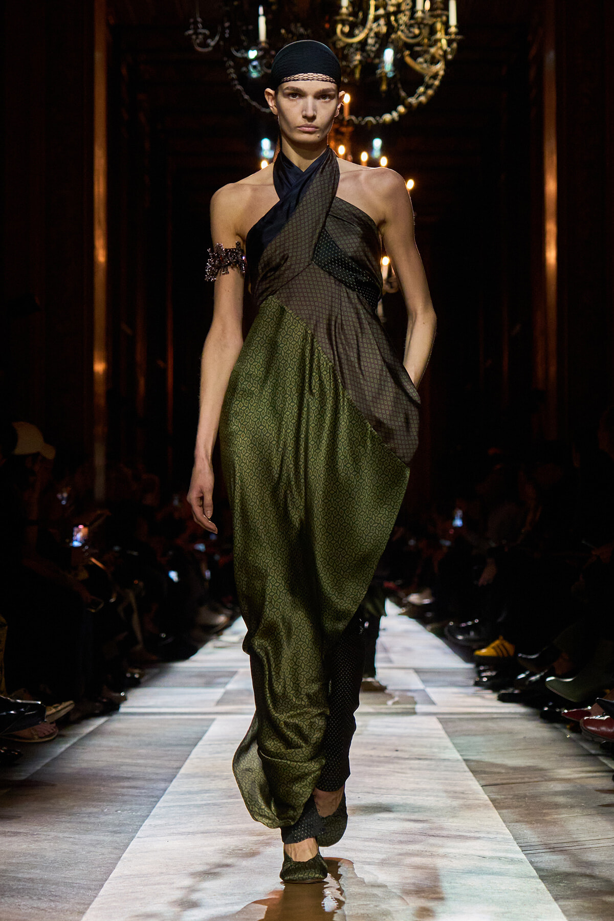 Model walks a fashion runway in a green geometric-patterned draped outfit with a halter-style top and black headscarf, chandeliers glowing in the background.