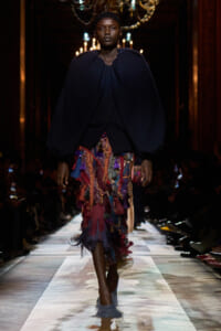 Model walking the runway in a voluminous navy cape over a multicolored tassel fringe skirt under chandelier lighting.