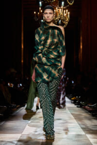Model walks a runway in a green plaid draped top and polka-dot pants, with chandeliers lit behind her in a fashion show setting.